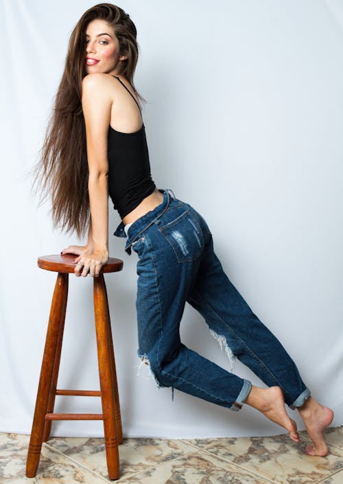Young Woman in Black Blouse and Distressed Jeans Leaning on a Stool