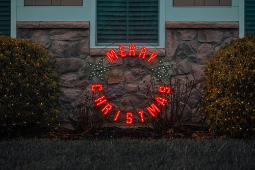 Free A vibrant 'Merry Christmas' wreath with glowing lights on a garden wall, creating a festive atmosphere. Stock Photo