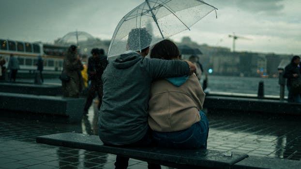 A couple sits close under an umbrella, embracing on a rainy city day.