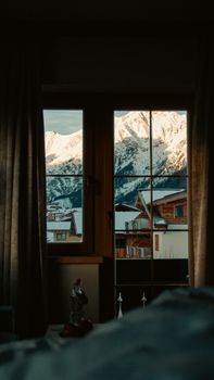 Cozy room view of snow-covered mountains and wooden cabins through a window.