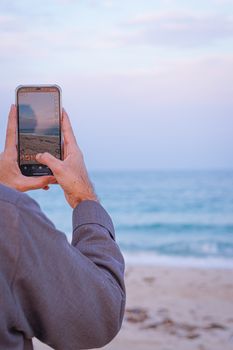 Person photographs serene beach scene with smartphone, capturing waves and sand.