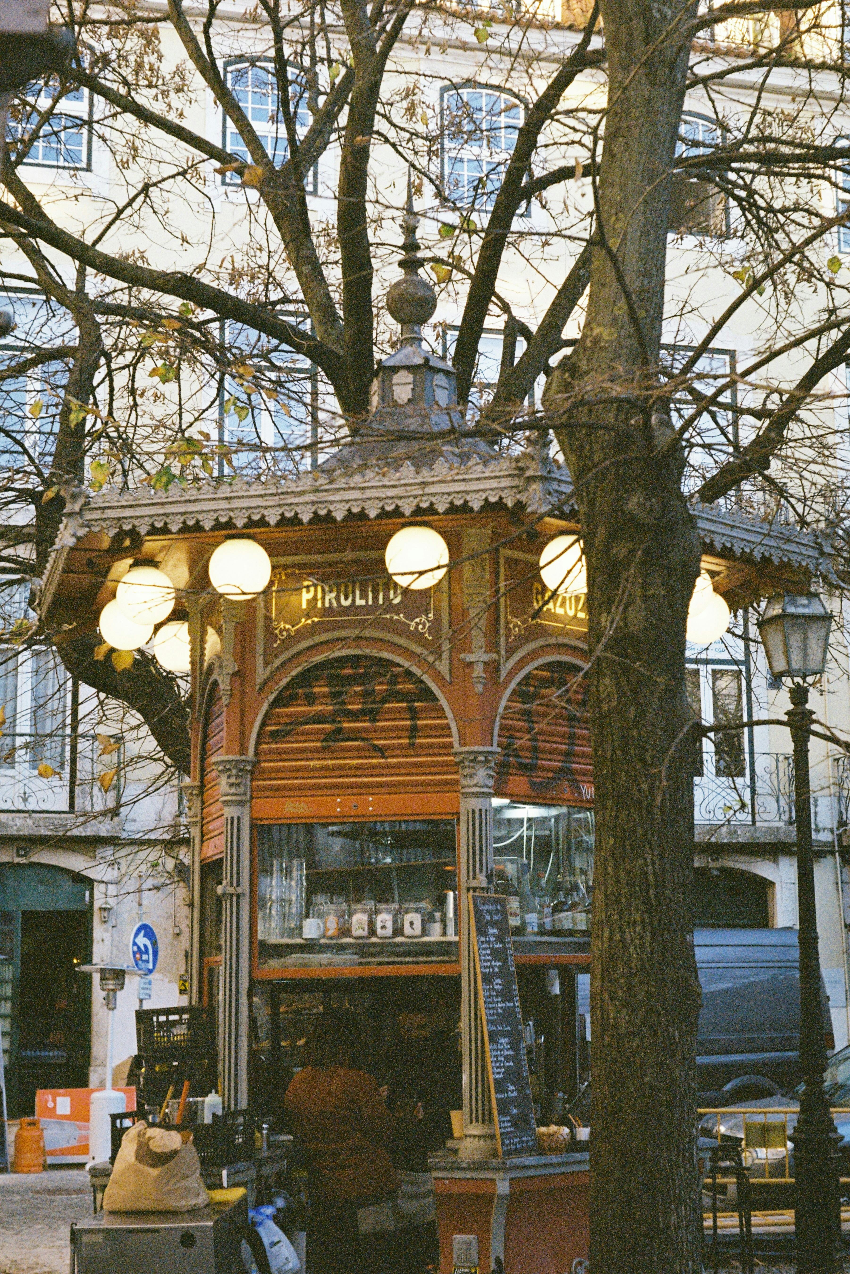 A vintage street food kiosk in Lisbon, surrounded by autumn trees and urban architecture.