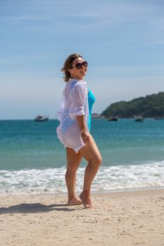Woman in beachwear strolling along a sunny shore with calm blue waters and boats in the distance.