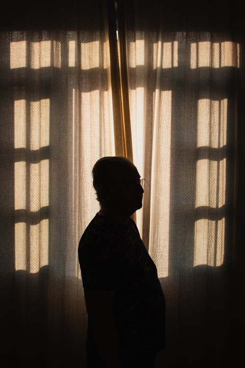 Side view of faceless man in glasses standing in room near window with closed curtains and looking away in soft daylight