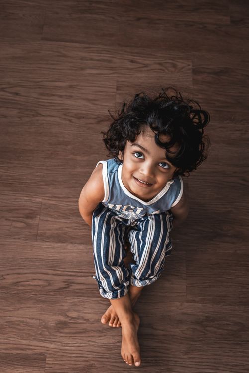 Free A charming child with curly hair smiling while sitting on a wooden floor indoors. Stock Photo