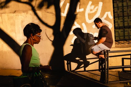 Free Side view of African American female in glasses walking down street with bag while ethnic man surfing web while sitting on fence in city in bright daylight Stock Photo