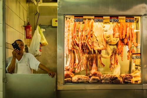 Busy ethnic butcher in white clothes speaking on phone and looking away near stand with meat in local market in soft light