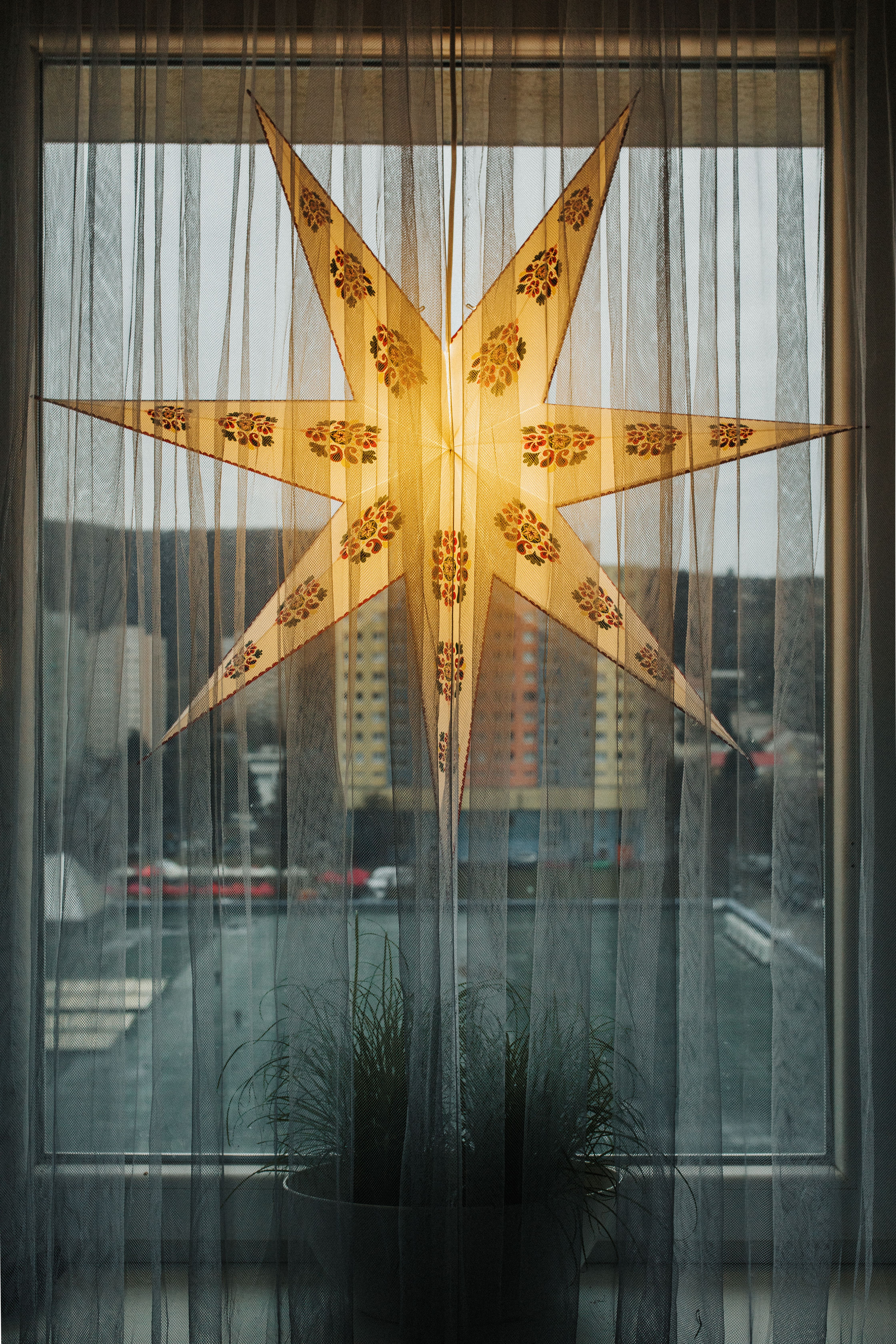 A glowing star-shaped decoration hanging on a window curtain with cityscape view.