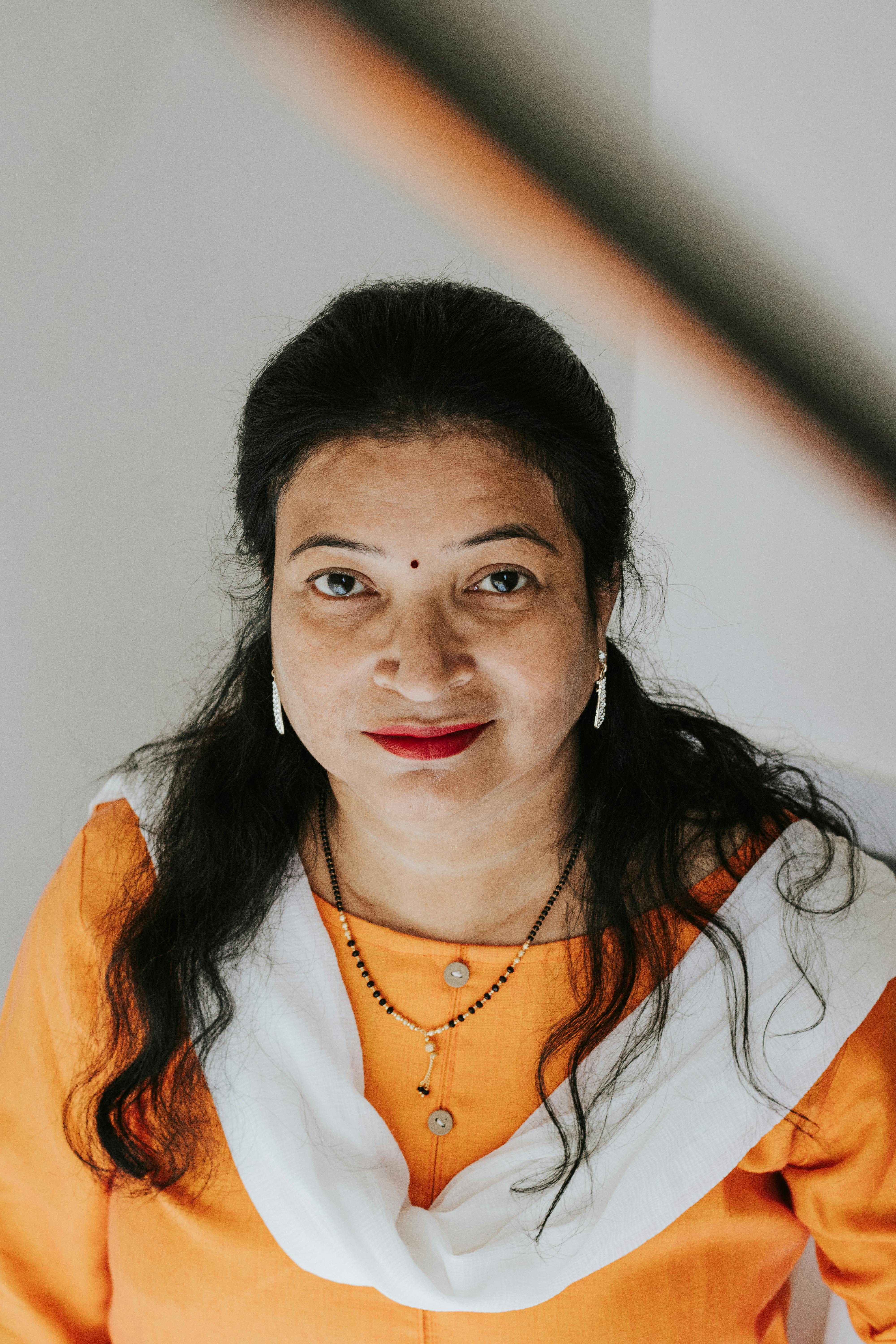 A woman in traditional orange attire smiles confidently indoors, captured in a front-facing portrait.