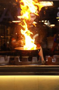 A chef creates a dramatic flare-up while cooking with a pan in a bustling kitchen.