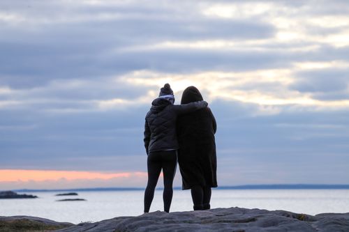 Free Two women in winter coats embracing by the seaside during sunset, depicting friendship and tranquility. Stock Photo