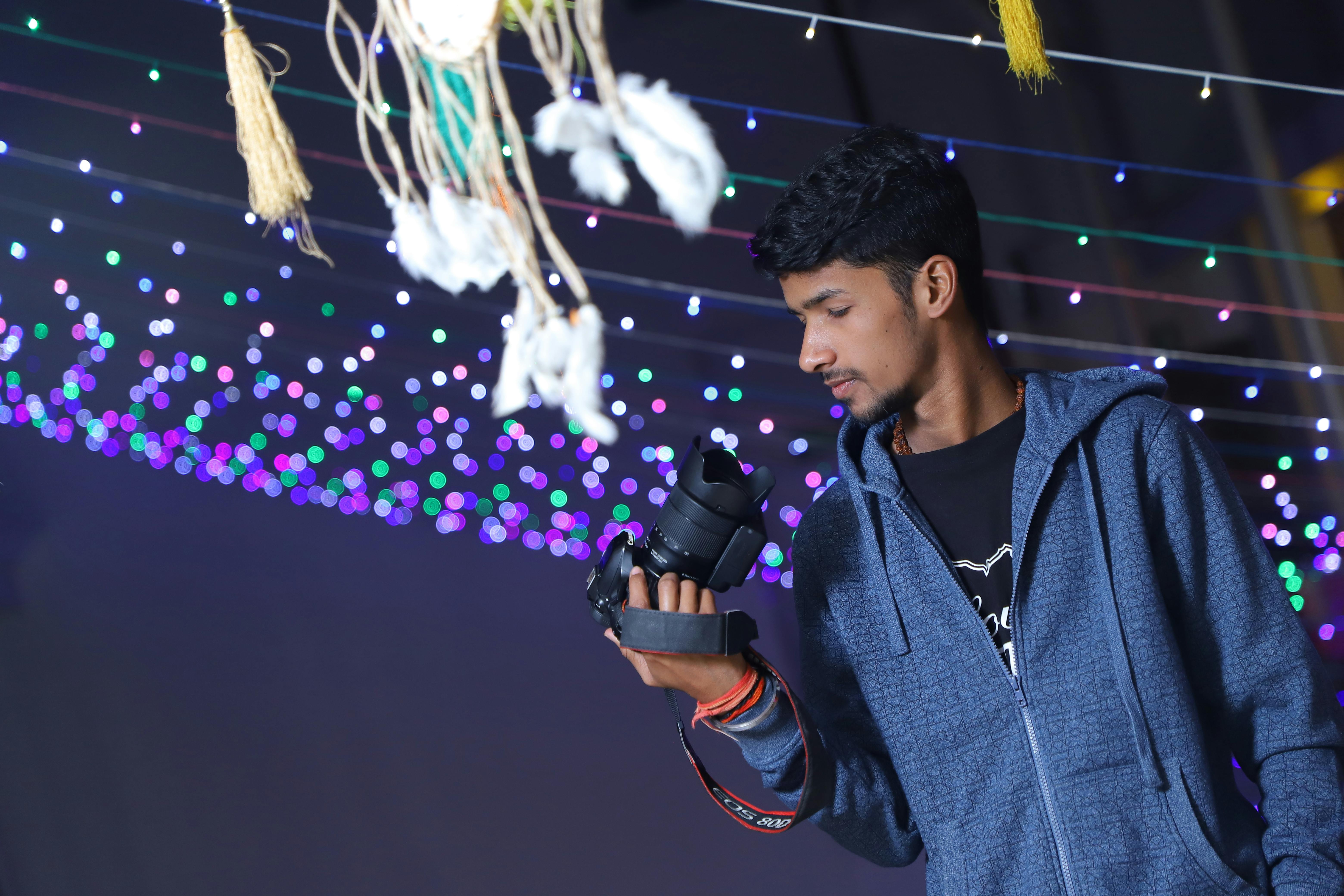 A young adult man in a hoodie holds a DSLR camera surrounded by colorful bokeh lights.