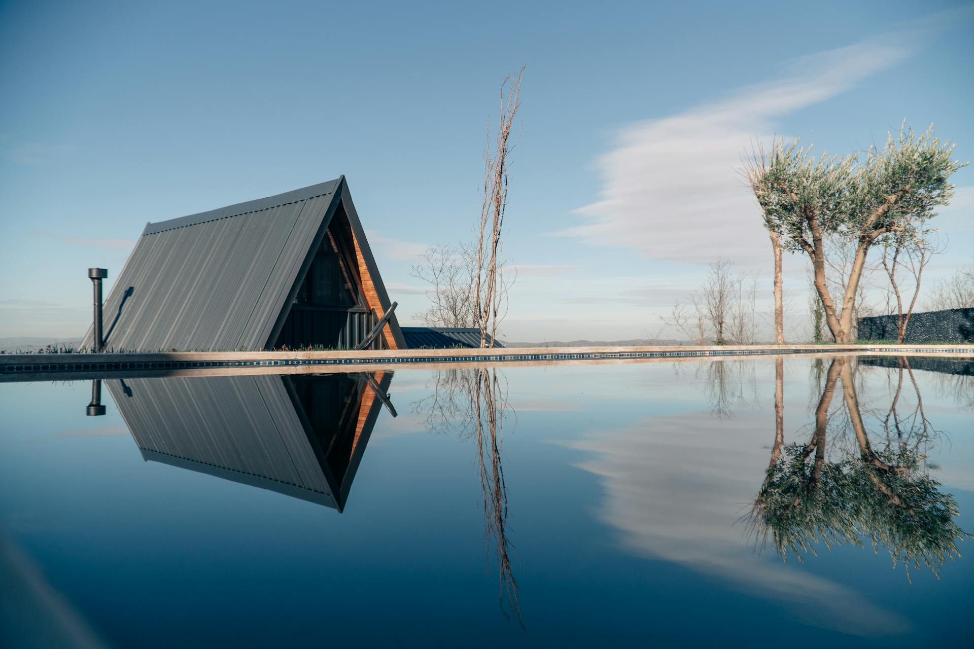 A sleek A-frame cabin with a metal roof reflecting in a serene swimming pool under a clear blue sky.