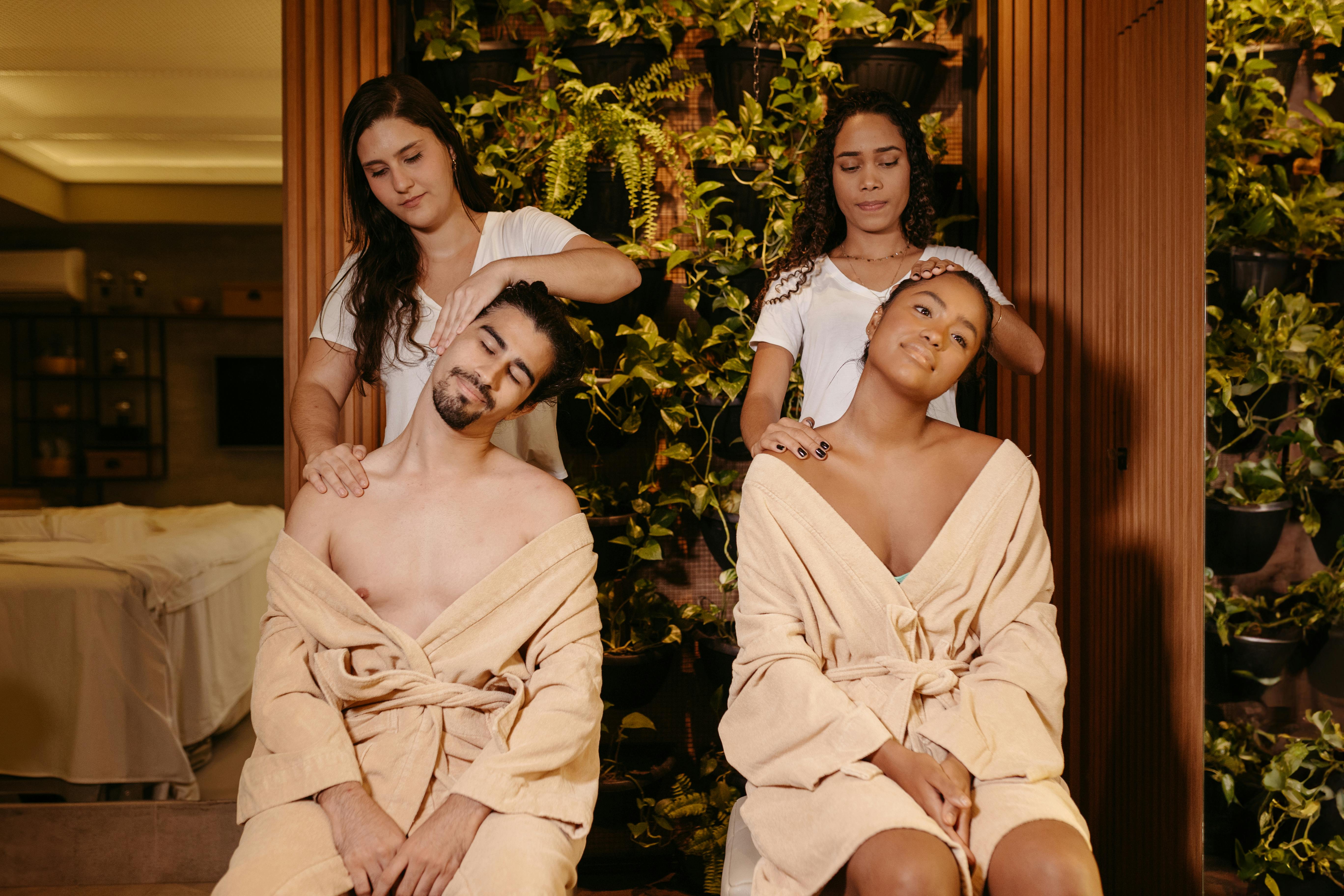 Two people in beige robes receive neck and shoulder massages from two practitioners in a lush, indoor plant setting.