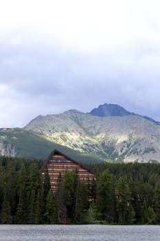 A serene mountain lodge nestled in lush forest by a tranquil alpine lake, under a cloudy sky.