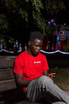 A young black man seated on a bench using a smartphone outdoors at night.