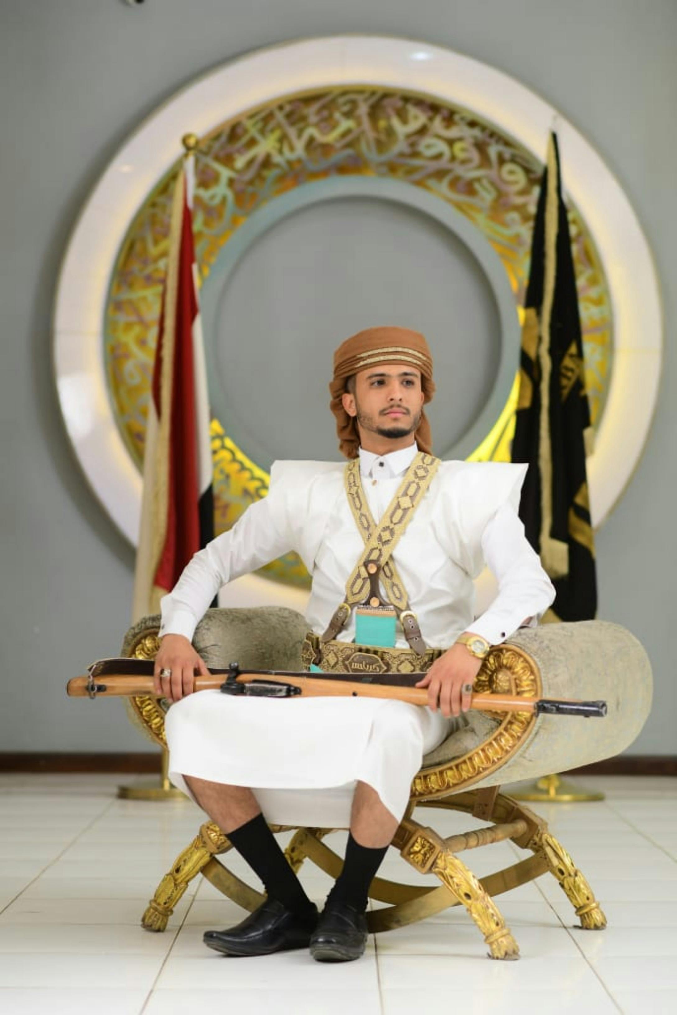 A man in traditional Arabian clothing, seated elegantly with cultural flags in the background.