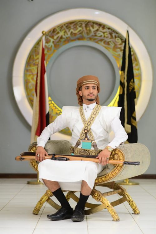 Free A man in traditional Arabian clothing, seated elegantly with cultural flags in the background. Stock Photo