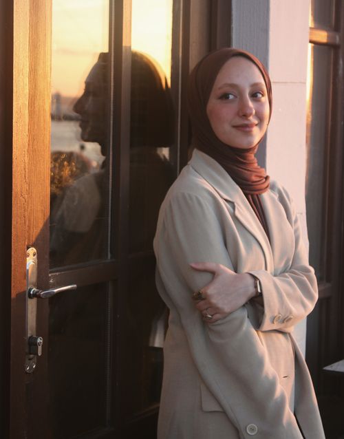 Free A fashionable woman in a coat and headscarf smiling by a window reflecting sunset light. Stock Photo