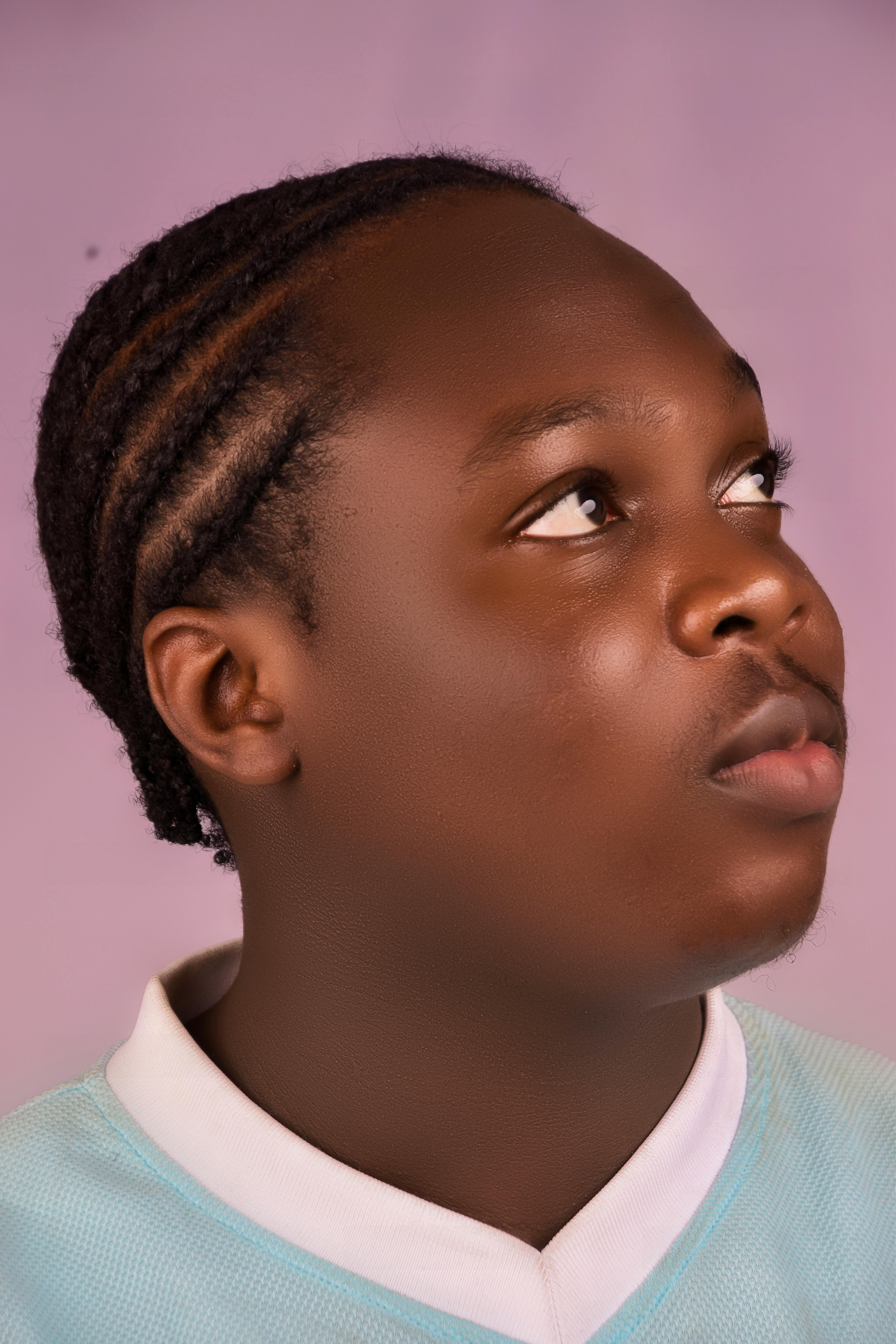 Studio portrait of an African teenage boy with braided hair looking up against a pink background.