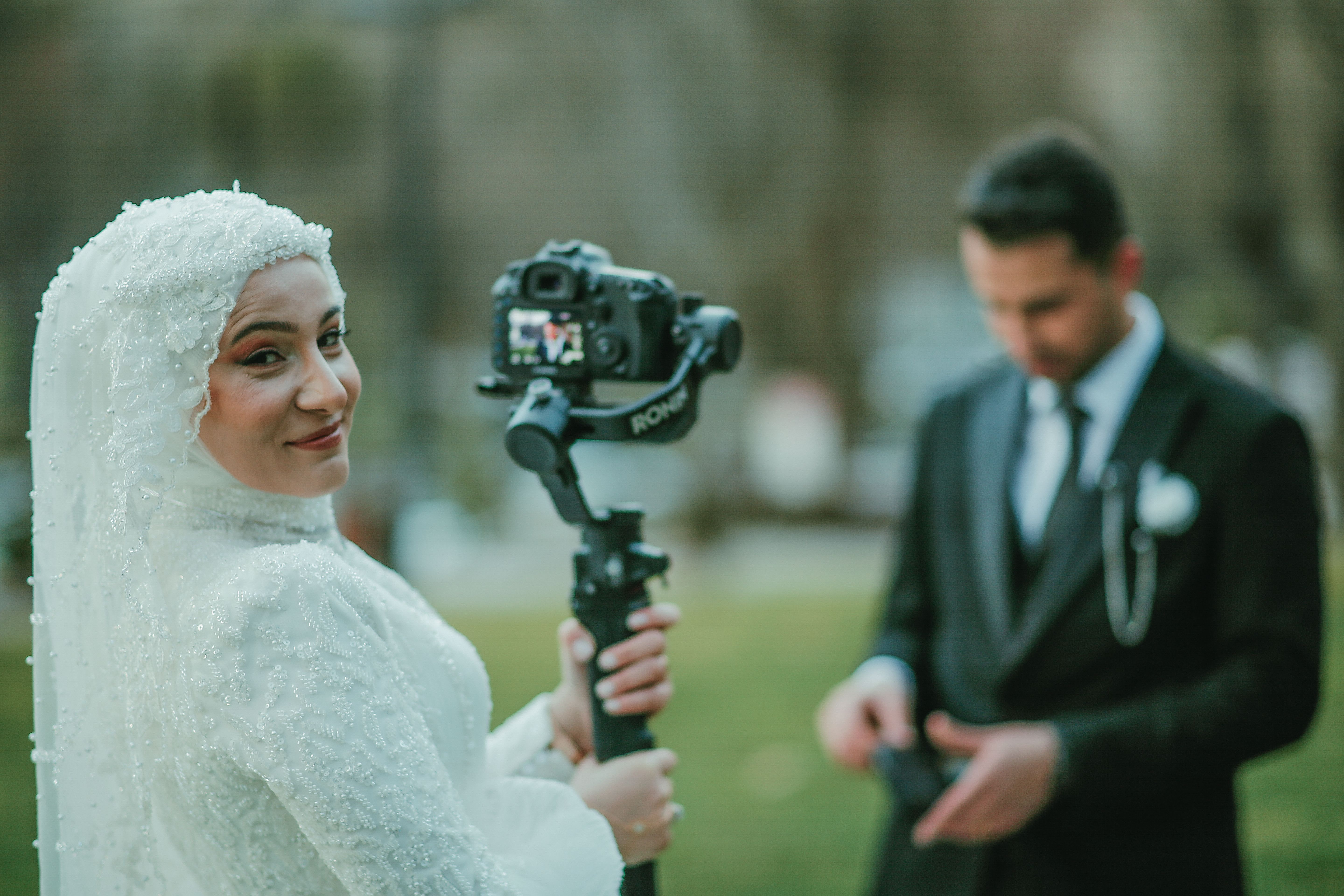 A bride in a white dress joyfully holding a camera next to her groom on their wedding day outdoors.