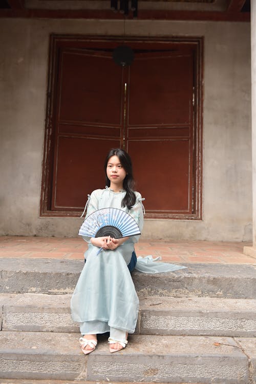 Young Woman in a Blue Ao Dai Dress Sitting on the Stairs Holding a Fan
