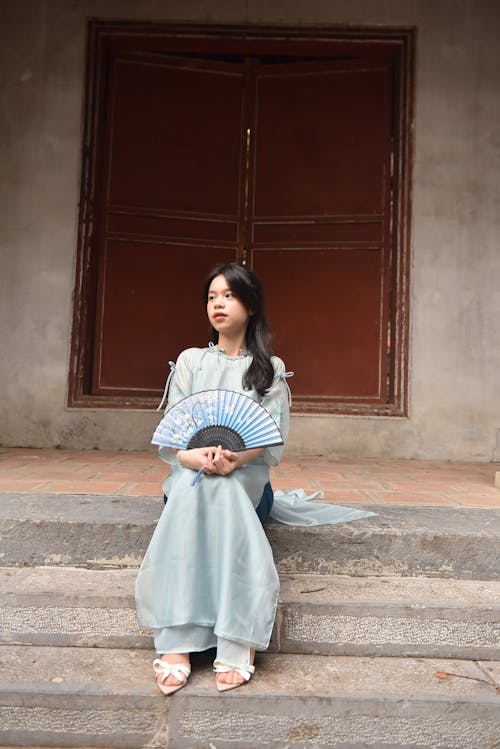 Young Model with Blue Paper Fan Wearing Traditional Vietnamese Dress Sitting on the Steps in Front of the Door
