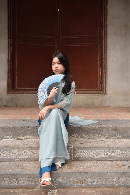 Young Model in a Blue Ao Dai Tunic with a Paper Fan Sitting on the Steps