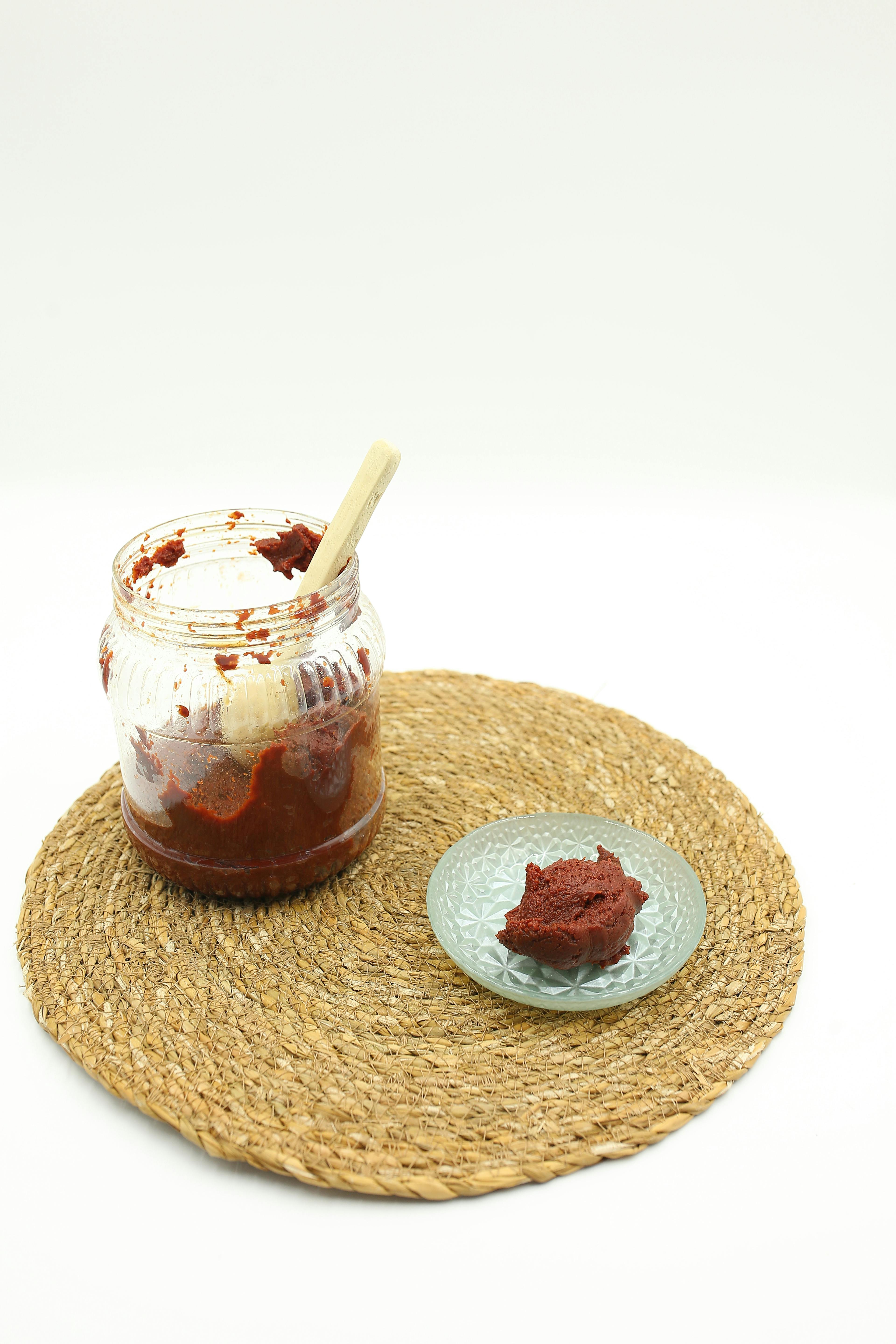 A jar and plate with red paste on a woven mat against a white background.