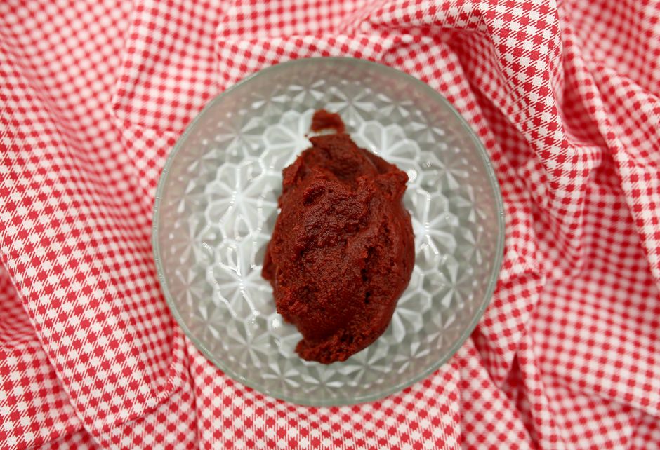 A small glass bowl of dark red tomato paste next to a halved tomato, a tube of paste, and a can, showing the thick concentrated texture compared to fresh tomato flesh