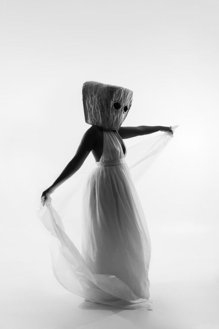 Artistic black and white portrait of a woman in a flowing dress, wearing a wooden mask.