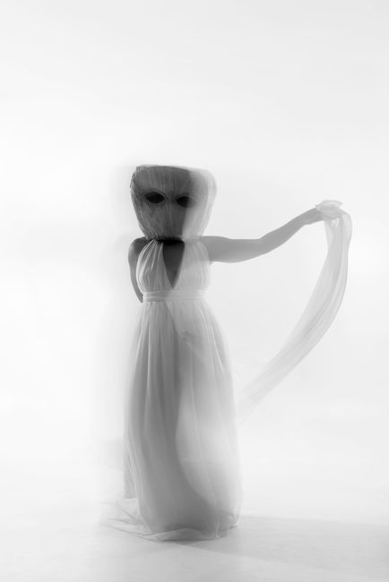 An artistic black and white photo of a woman in a mask and dress, evoking mystery and movement.