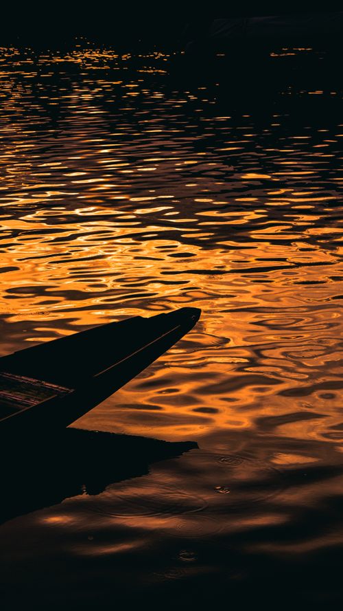 Free A silhouetted boat against a golden sunset reflecting on calm rippling water. Stock Photo