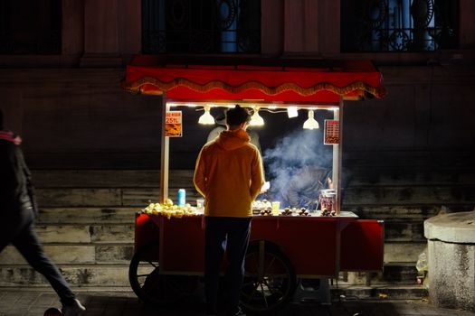 A street vendor stands at a well-lit cart selling food at night in an urban setting.