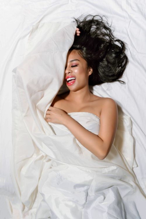 Free A joyful woman lying comfortably in bed wrapped in white sheets, radiating happiness. Stock Photo
