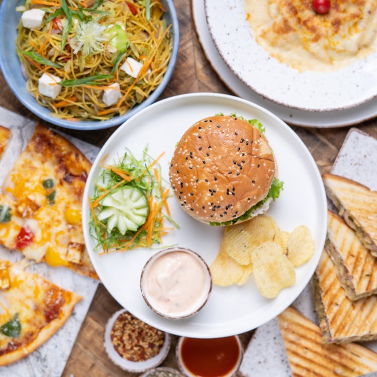 Plates And Bowls With A Burger, Sandwiches, And Pizza On A Table