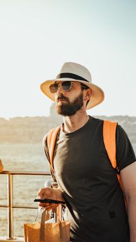 Man in sunglasses and hat with backpack by the sea during summer sunrise.