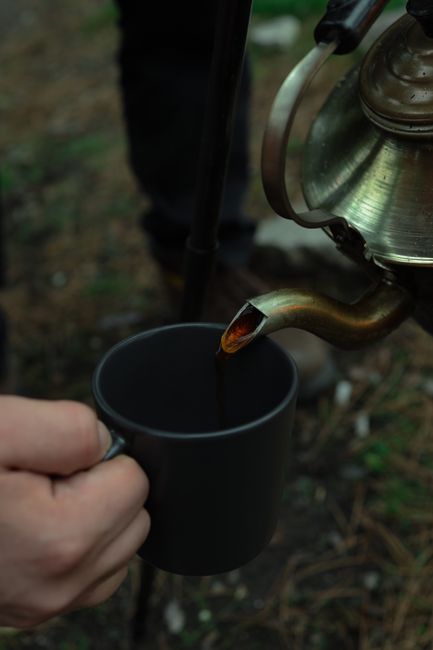 Outdoor tea being poured into a black camping mug from a kettle.