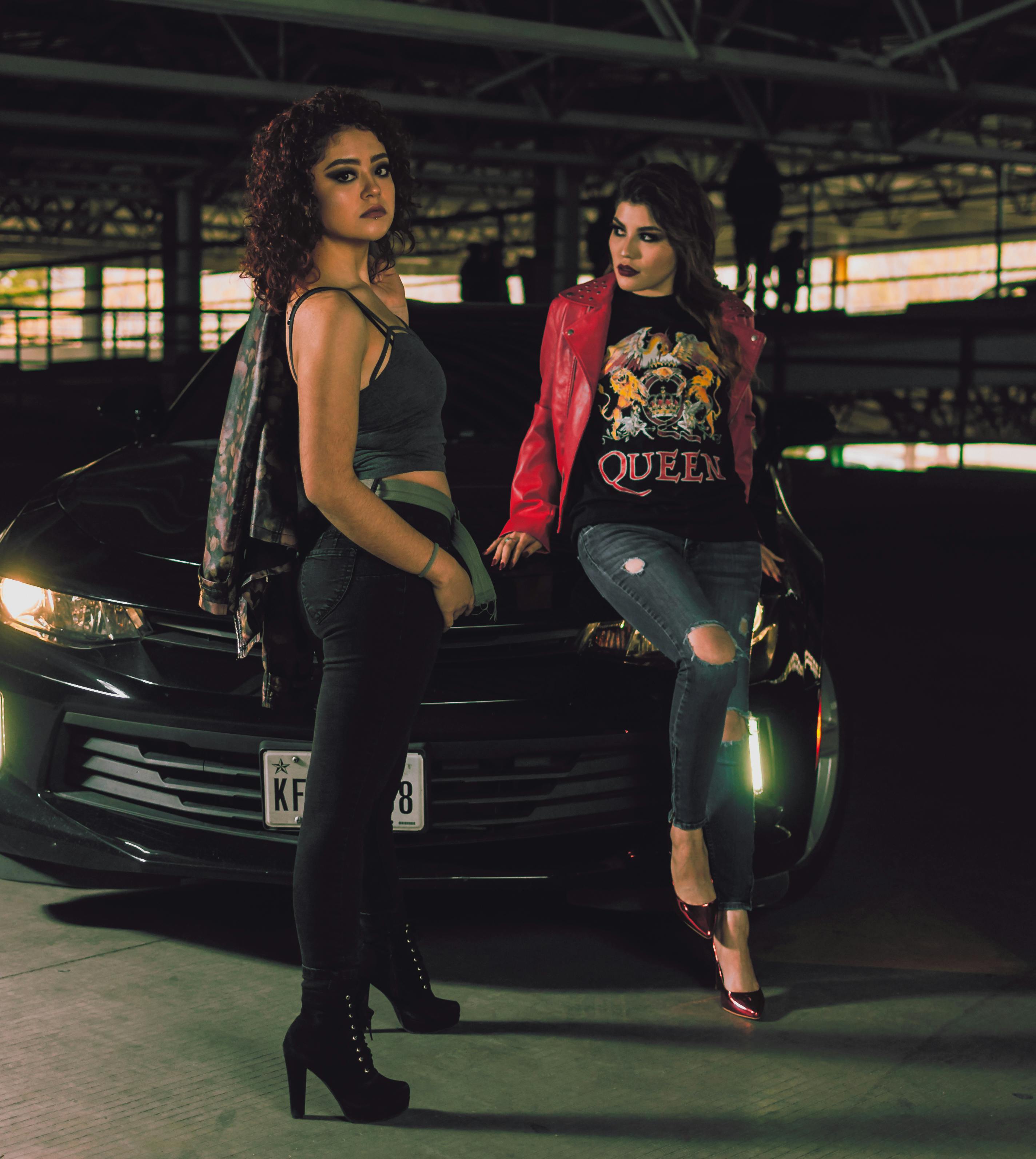 在光线昏暗的城市停车场，两名时尚女性自信地对着汽车摆姿势