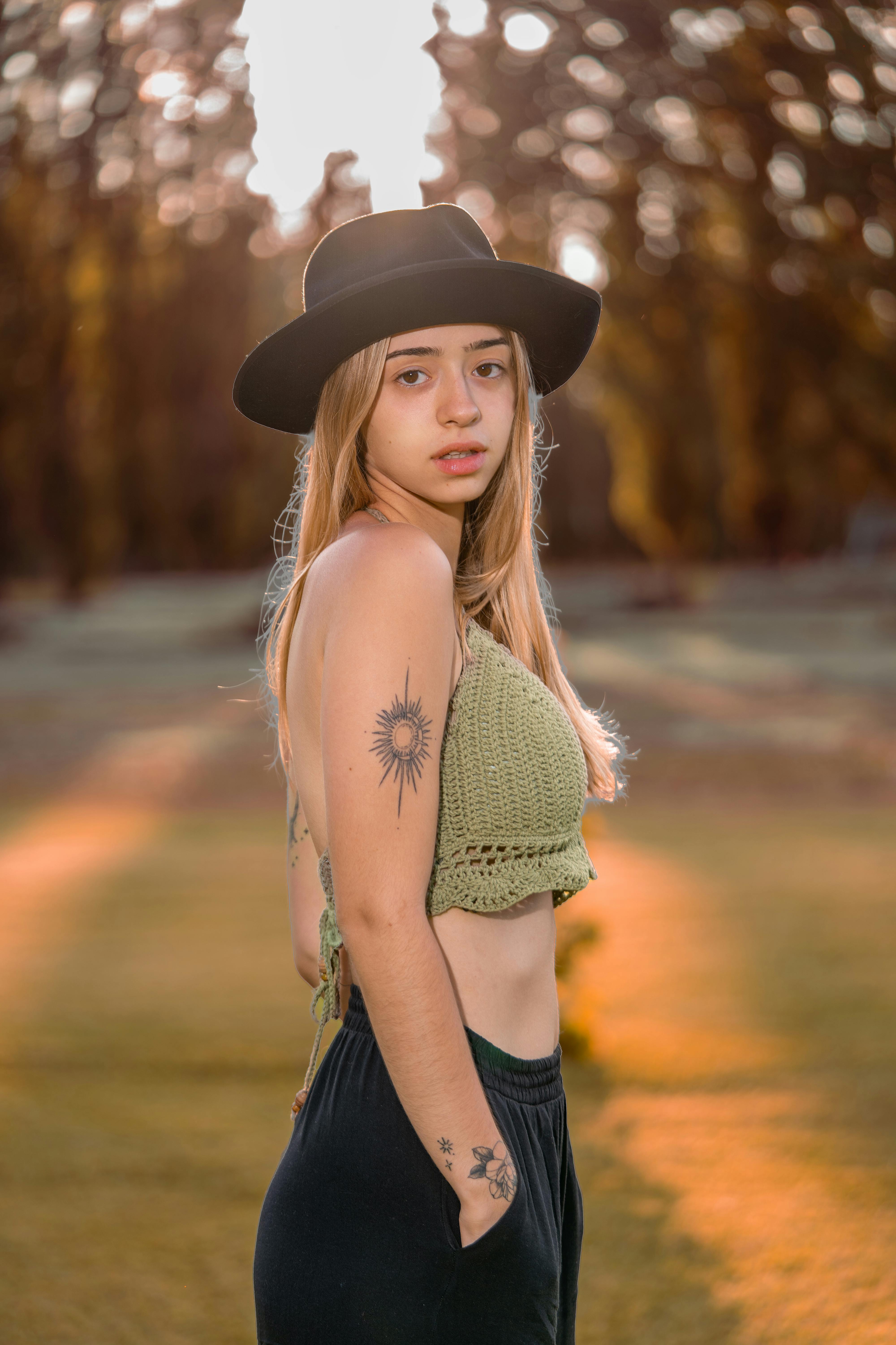 A fashionable young woman with tattoos poses outdoors in a sunlit autumn forest with a hat.