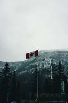 Canadian flag in Banff with snow-covered mountains and evergreen trees, capturing winter nature.