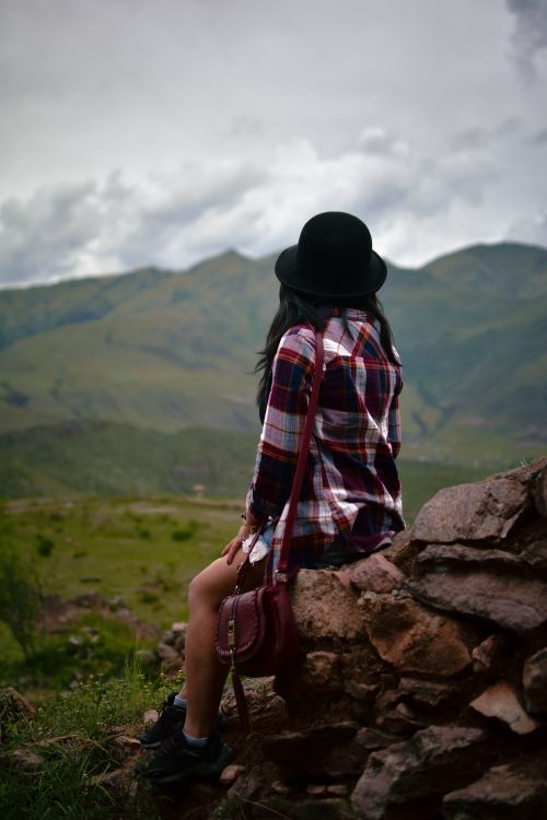 Free A woman in a plaid shirt sits calmly on rocks, gazing at a scenic mountain view. Stock Photo
