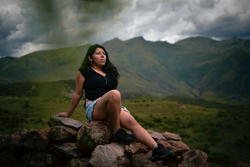 Free Woman sits on rocks against a breathtaking mountainous backdrop under dramatic skies. Stock Photo