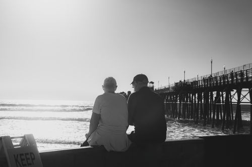 Free Two adults sit by a seaside pier, enjoying a calm sunset in black and white. Stock Photo