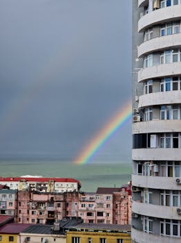 A striking rainbow arcs over a seaside cityscape against a backdrop of cloudy skies.