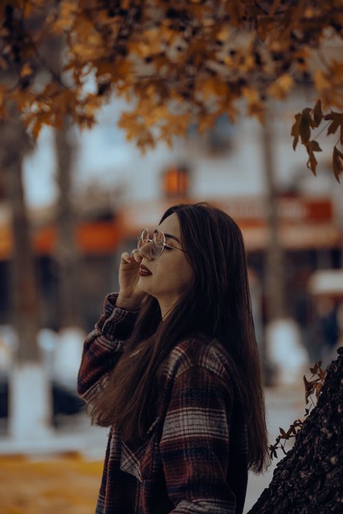 Free Side view of a young woman standing near a tree wearing eyeglasses and a checkered jacket in a fall setting. Stock Photo