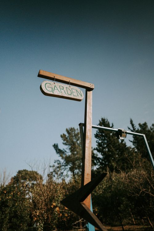 Free A rustic wooden garden sign stands against a clear blue sky with trees in the background. Stock Photo