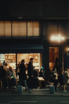 Nighttime urban street scene with people passing by an illuminated bar.