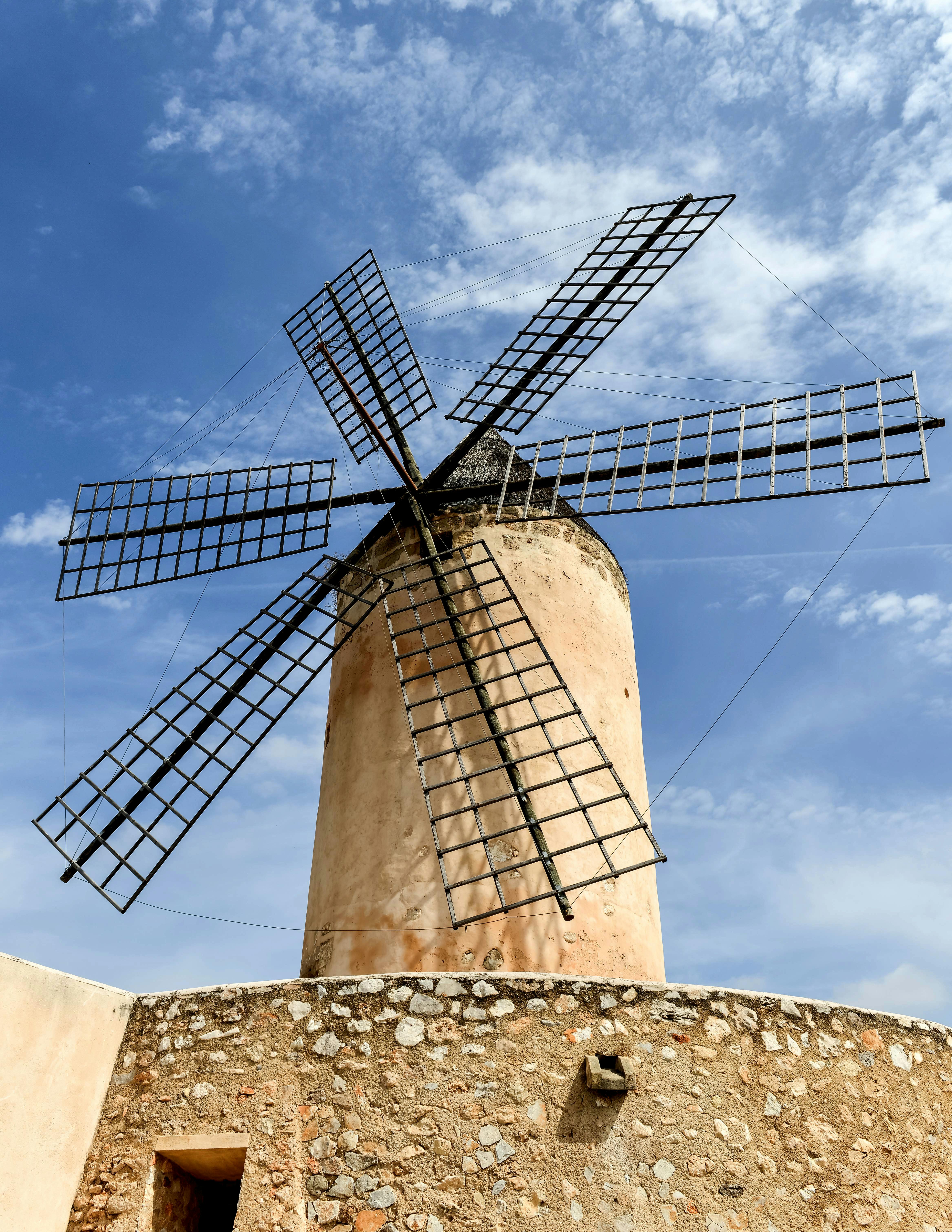 A stone windmill with metallic arms stands tall under a bright blue sky in rural Spain.