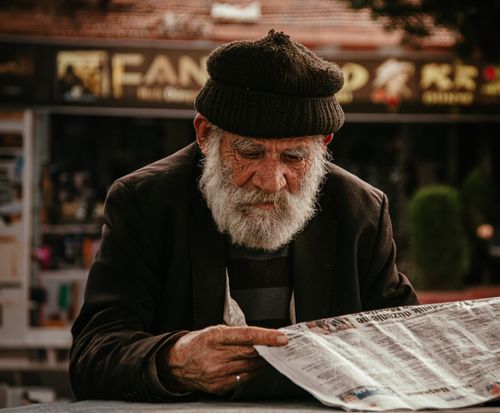 Free Senior man in woolen cap reading a newspaper on city streets, captured in a candid street photography style. Stock Photo